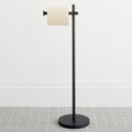 A freestanding toilet paper holder with a black finish and a modern design, featuring a cylindrical holder with a paper roll on it, placed on a tiled floor against a white wall.